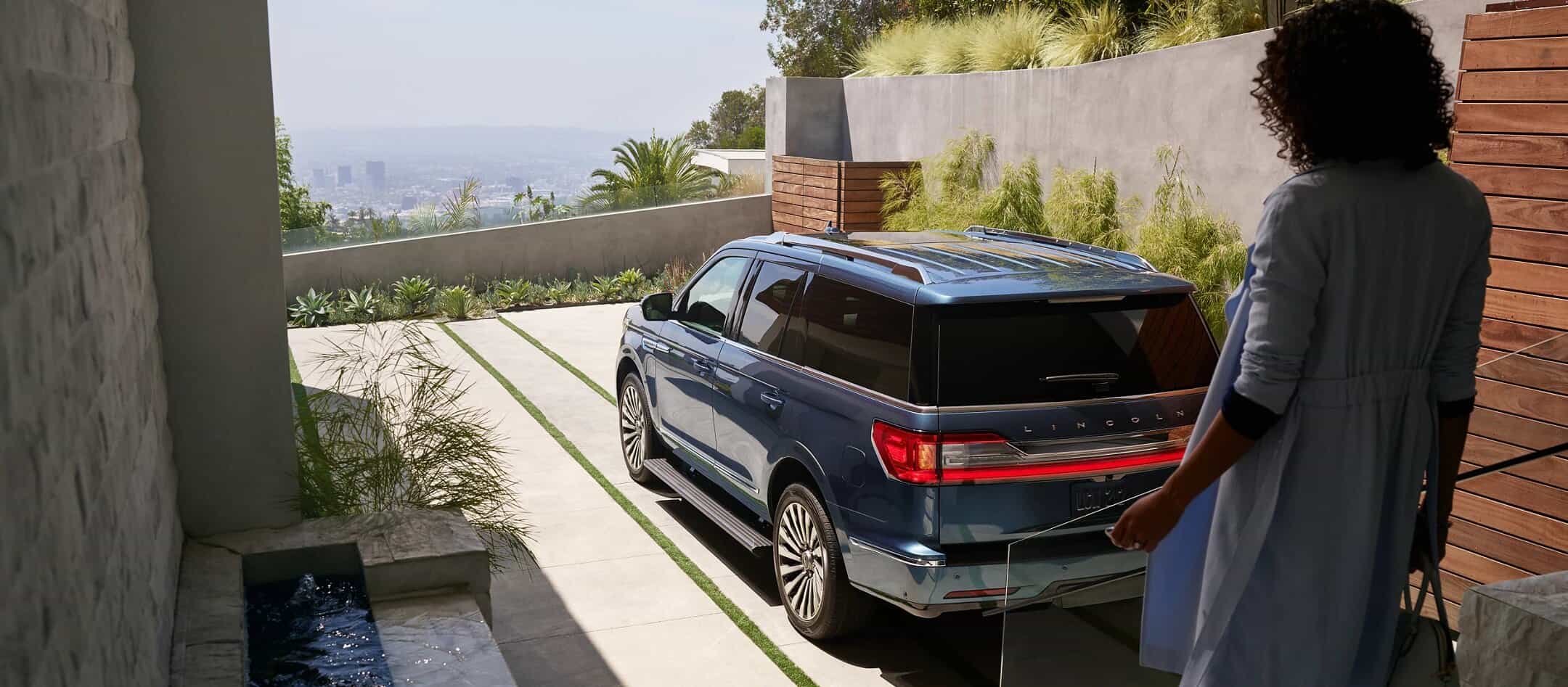 Person approaching a blue SUV parked in a modern driveway with cityscape views.
