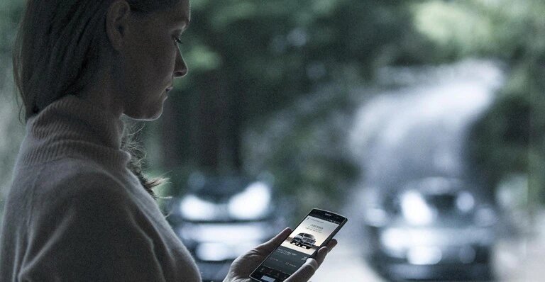 Woman using smartphone with car app, standing near blurred cars and trees in the background.