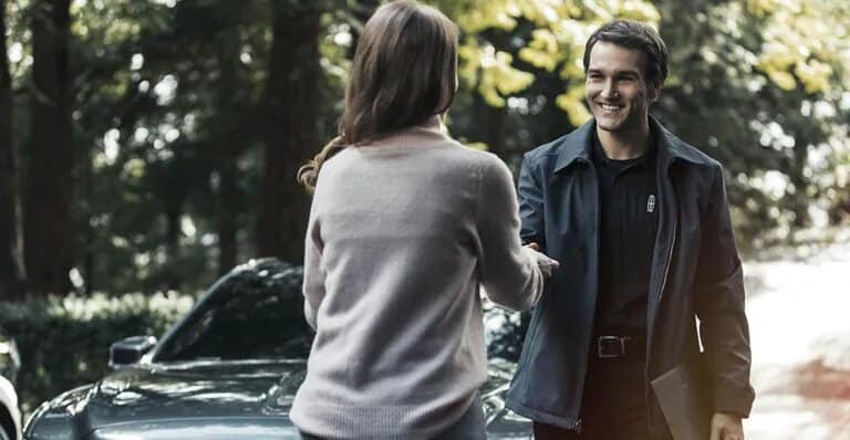 A woman and a man shake hands in front of a car, surrounded by outdoor greenery.