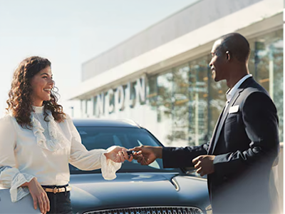 Woman receiving car keys from a man in front of a dealership, both smiling.
