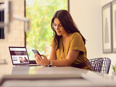 Woman in yellow dress using a smartphone at a table with a laptop, bright room background.